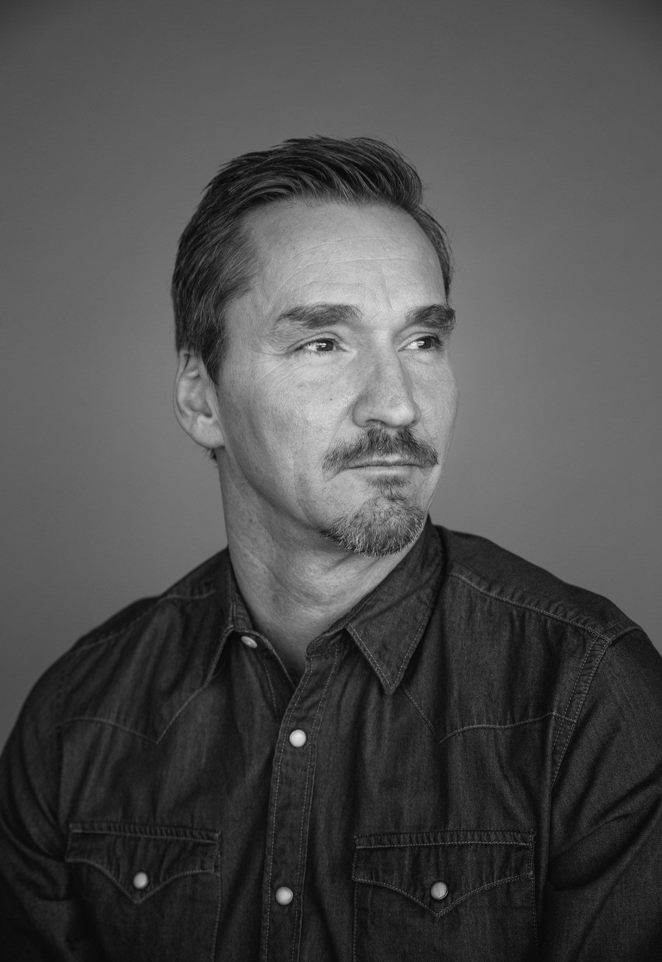 Black and white portrait of a man with dark hair and facial hair wearing a denim shirt