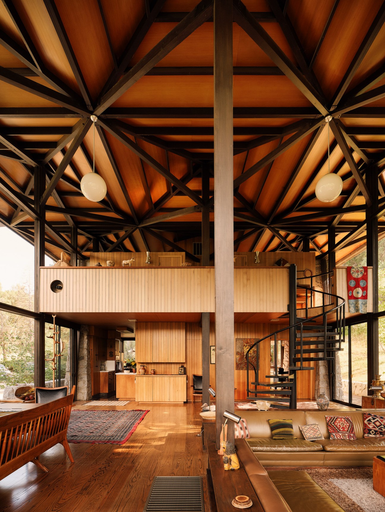 High-ceilinged modernist interior with exposed wooden beams, folk art textiles, and a spiral staircase.