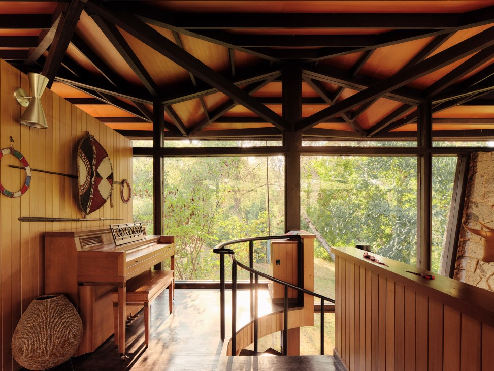 Elevated wooden landing with railings overlooking forest, featuring piano and Native American crafts beneath exposed beam ceiling.
