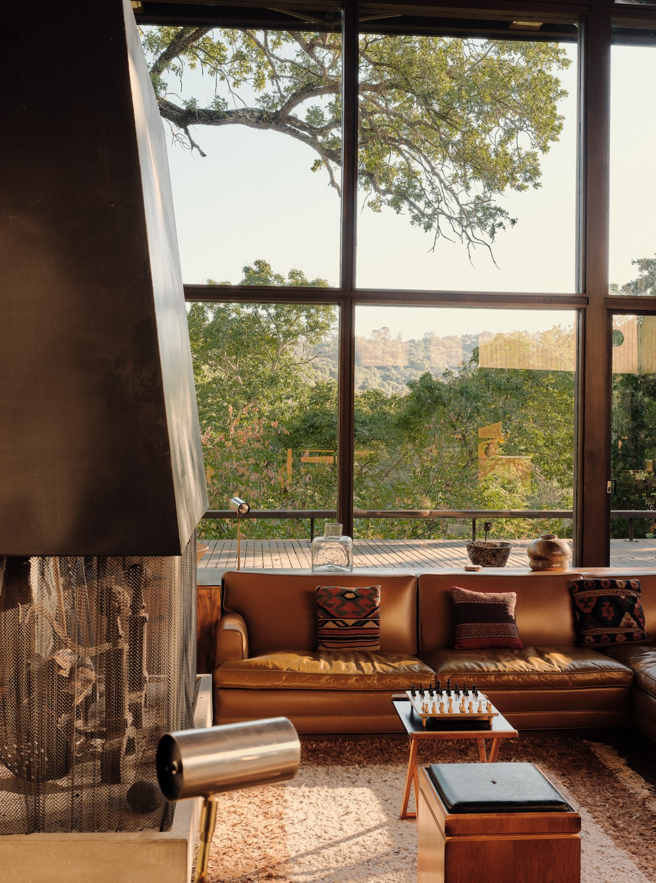 Sunken circular conversation area with brown leather seating and cushions around a central fireplace, framed by large windows overlooking natural landscape.