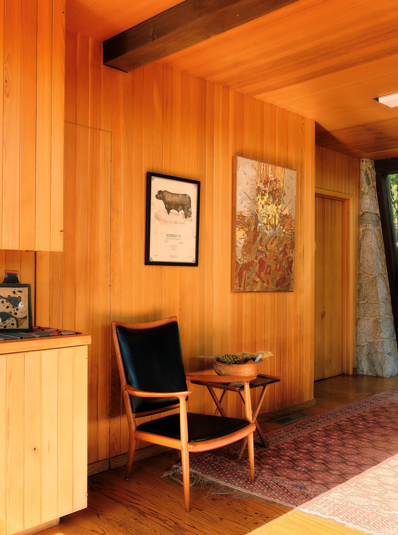 Wooden chair with curved armrests and black cushioned seat in mid-century modern interior with knotty pine walls.