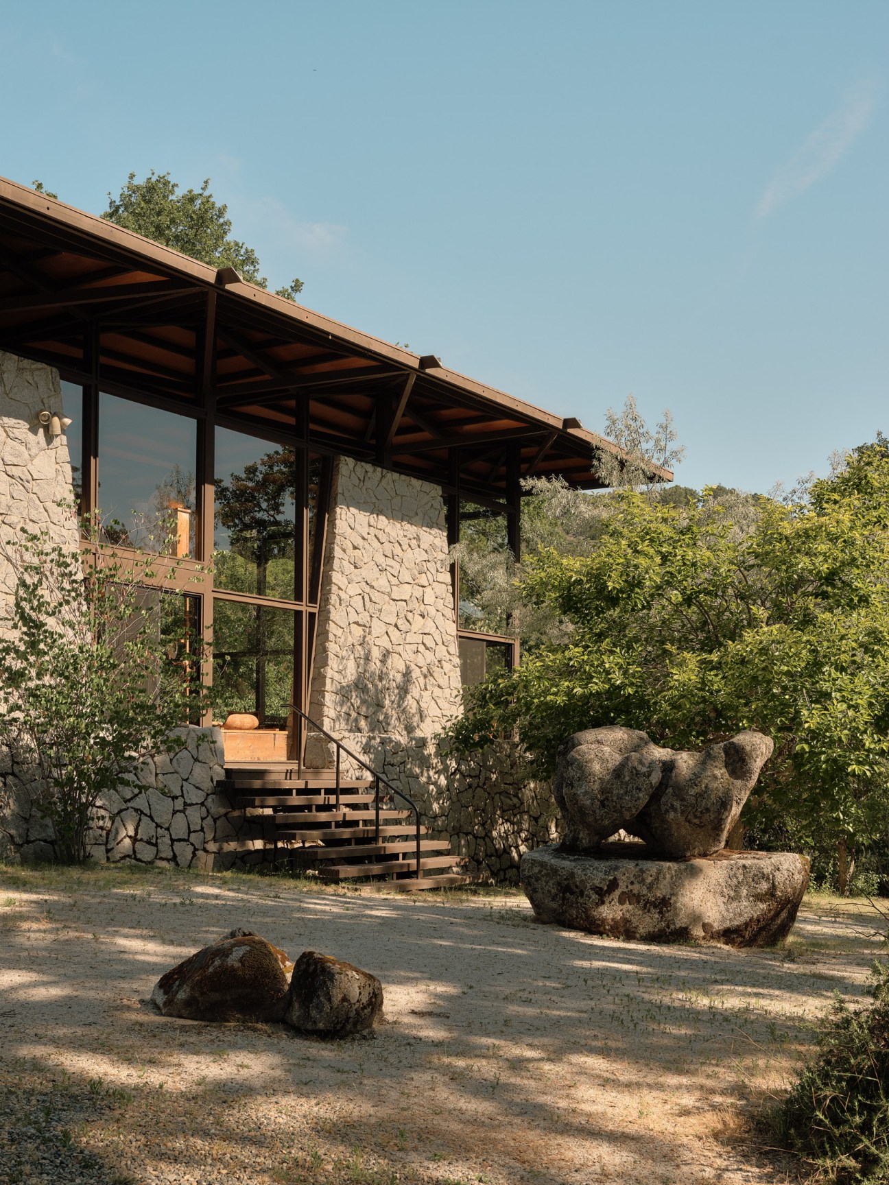 Modernist stone house with large wooden deck and sculptural granite boulder formation resembling a bear in foreground.