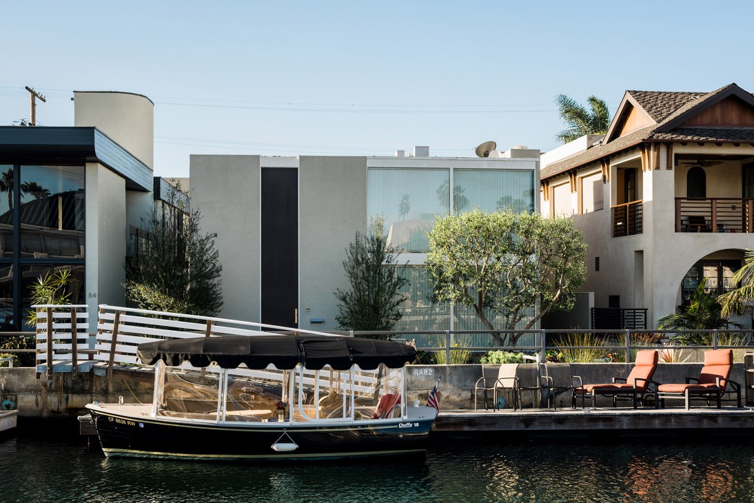 Modern mid-century house with large glass windows and olive trees sits on canal waterfront with boat moored in foreground.