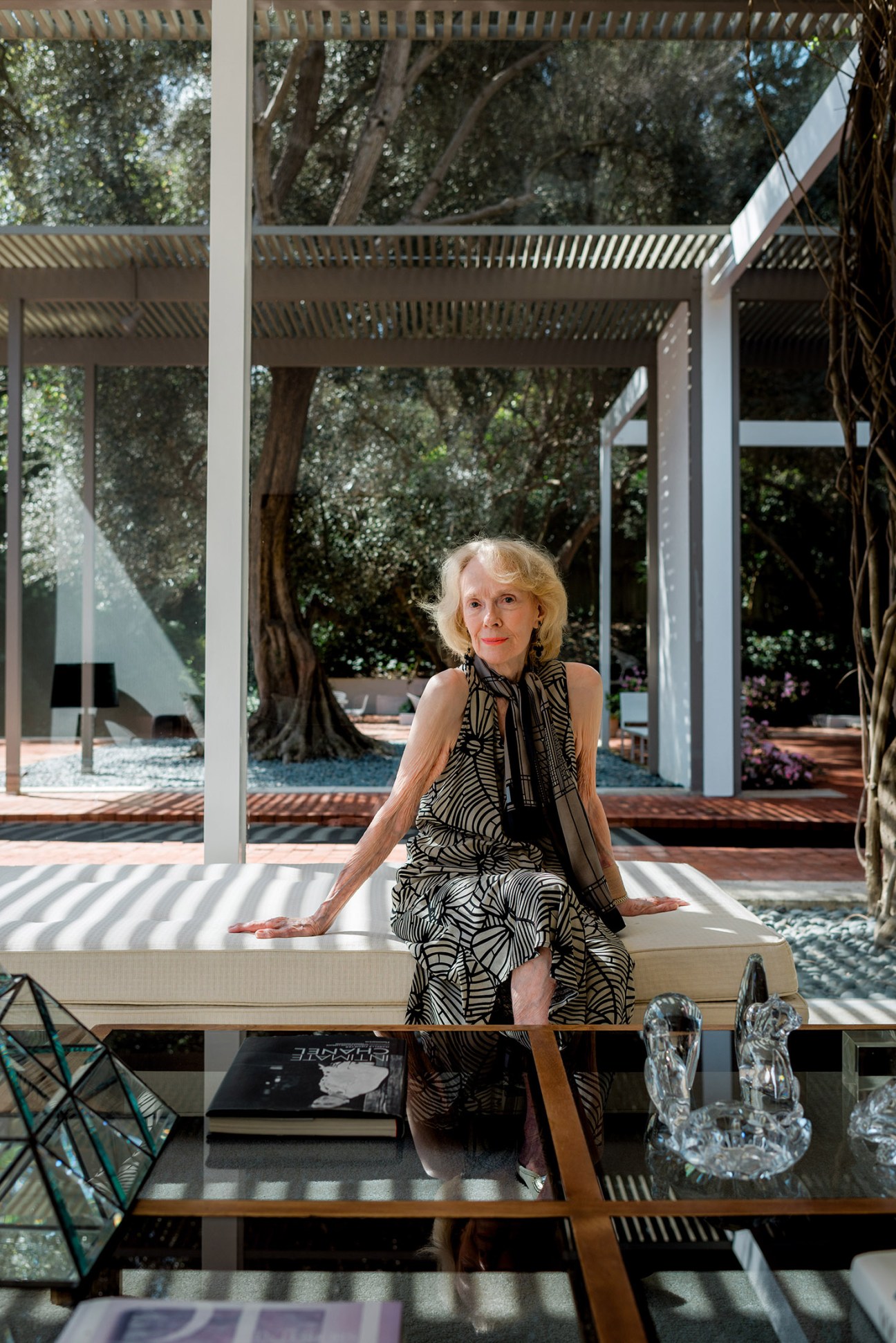 Laura Killingsworth sits on a bench inside her mid-century modern home, with large glass windows and olive trees visible behind her.