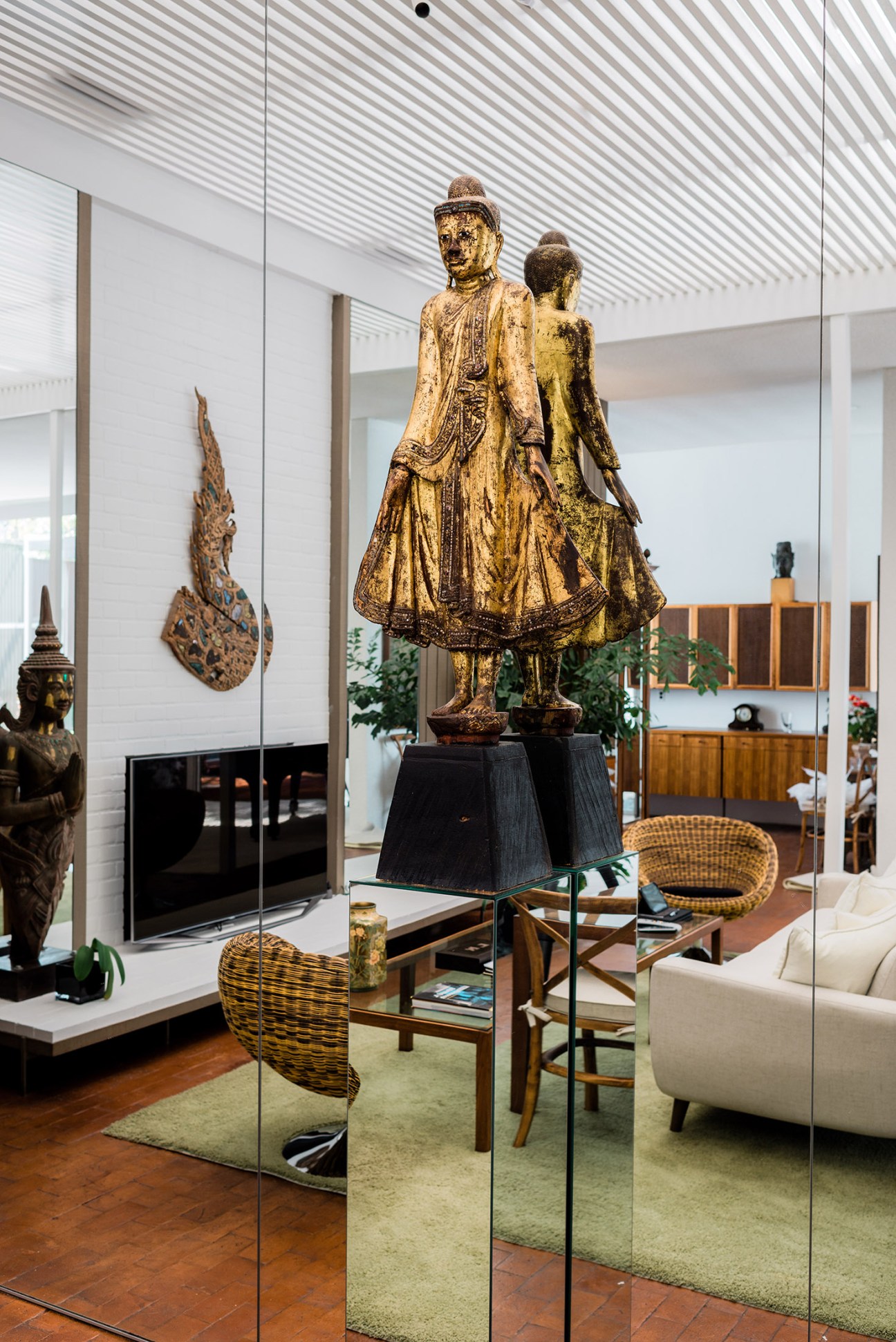Golden Buddhist statues and carved wooden masks displayed in a modernist interior with glass walls and mid-century furniture.
