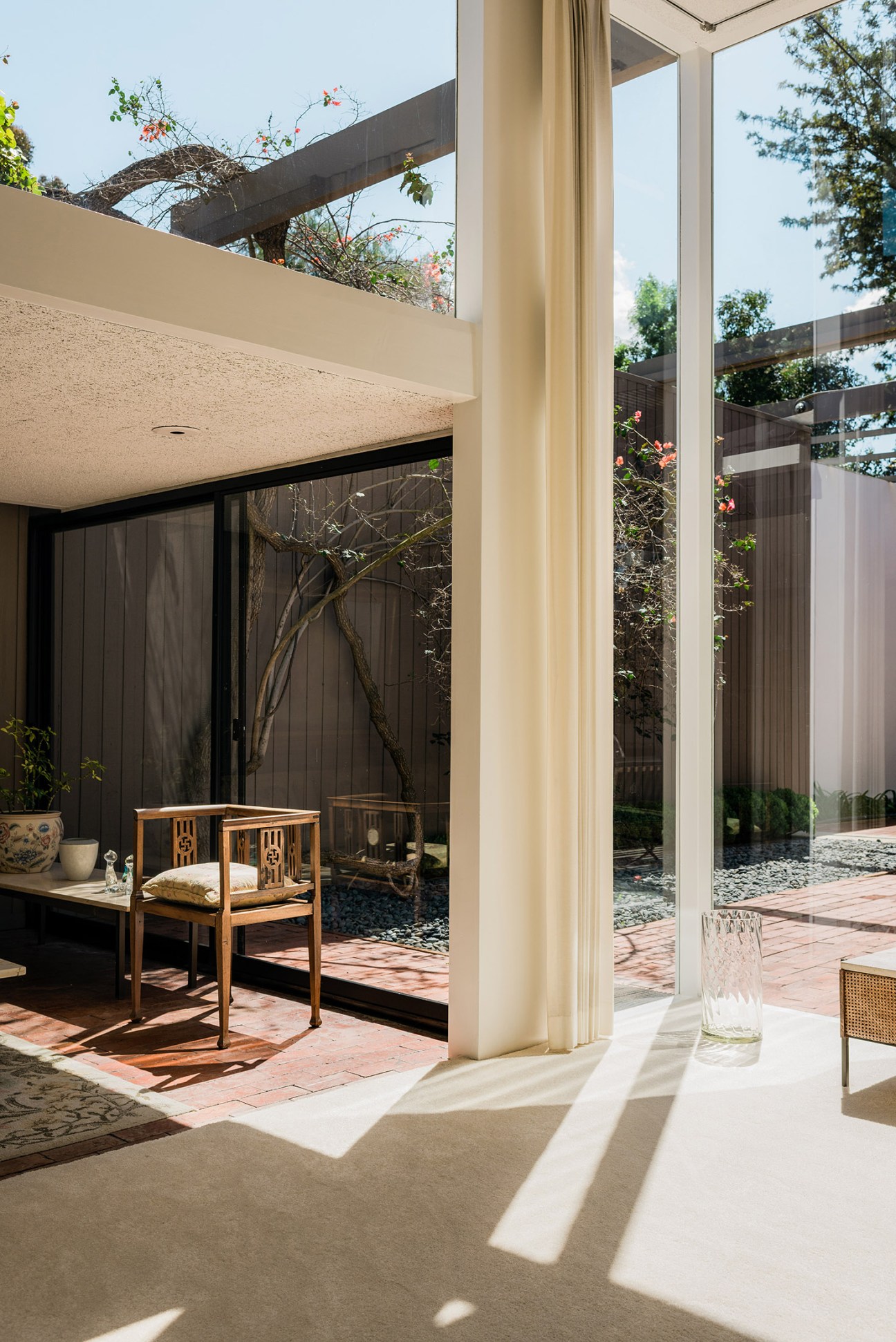 Modernist interior with floor-to-ceiling glass doors opening to a patio, showing indoor-outdoor space with wooden furniture and architectural details.