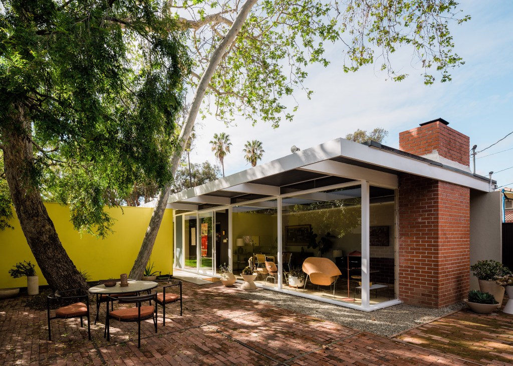 Mid-century modern house with brick and glass facade, white overhang, yellow wall, and mature tree in front patio.