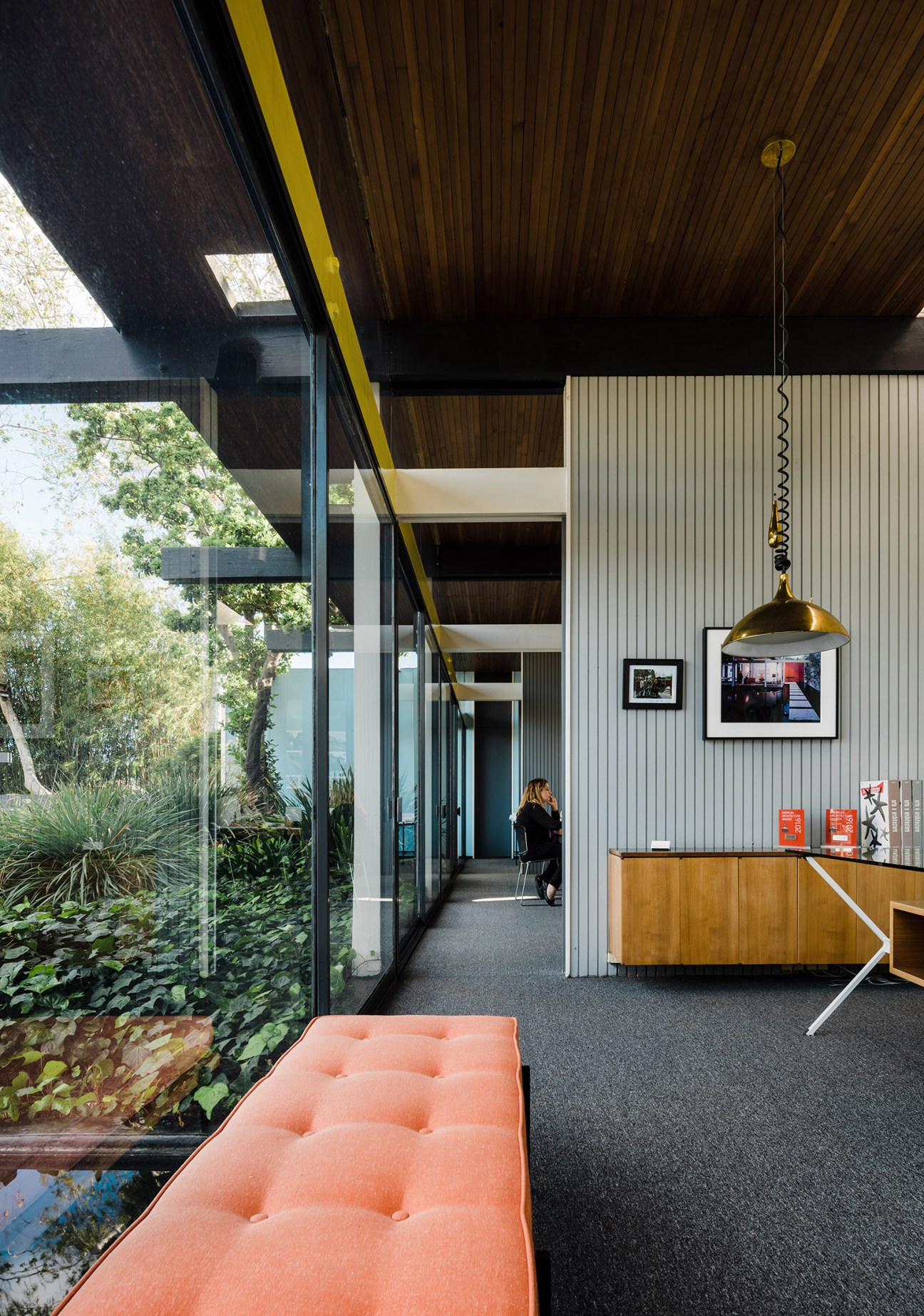 Interior of a modernist office with floor-to-ceiling windows, wooden ceiling, coral bench, and brass pendant light.
