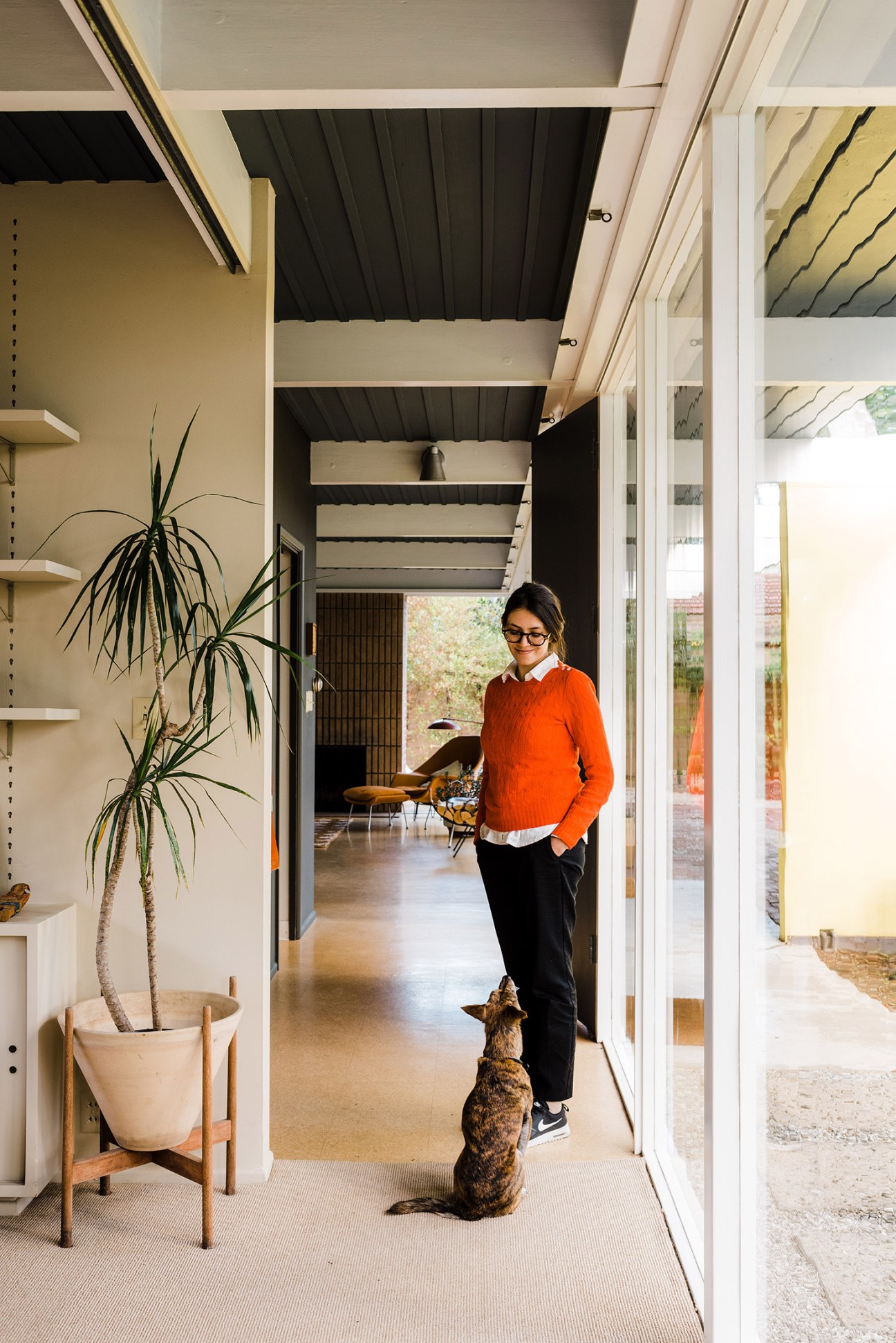 Woman in orange sweater walking with dog through modernist hallway with exposed beams and glass doors.