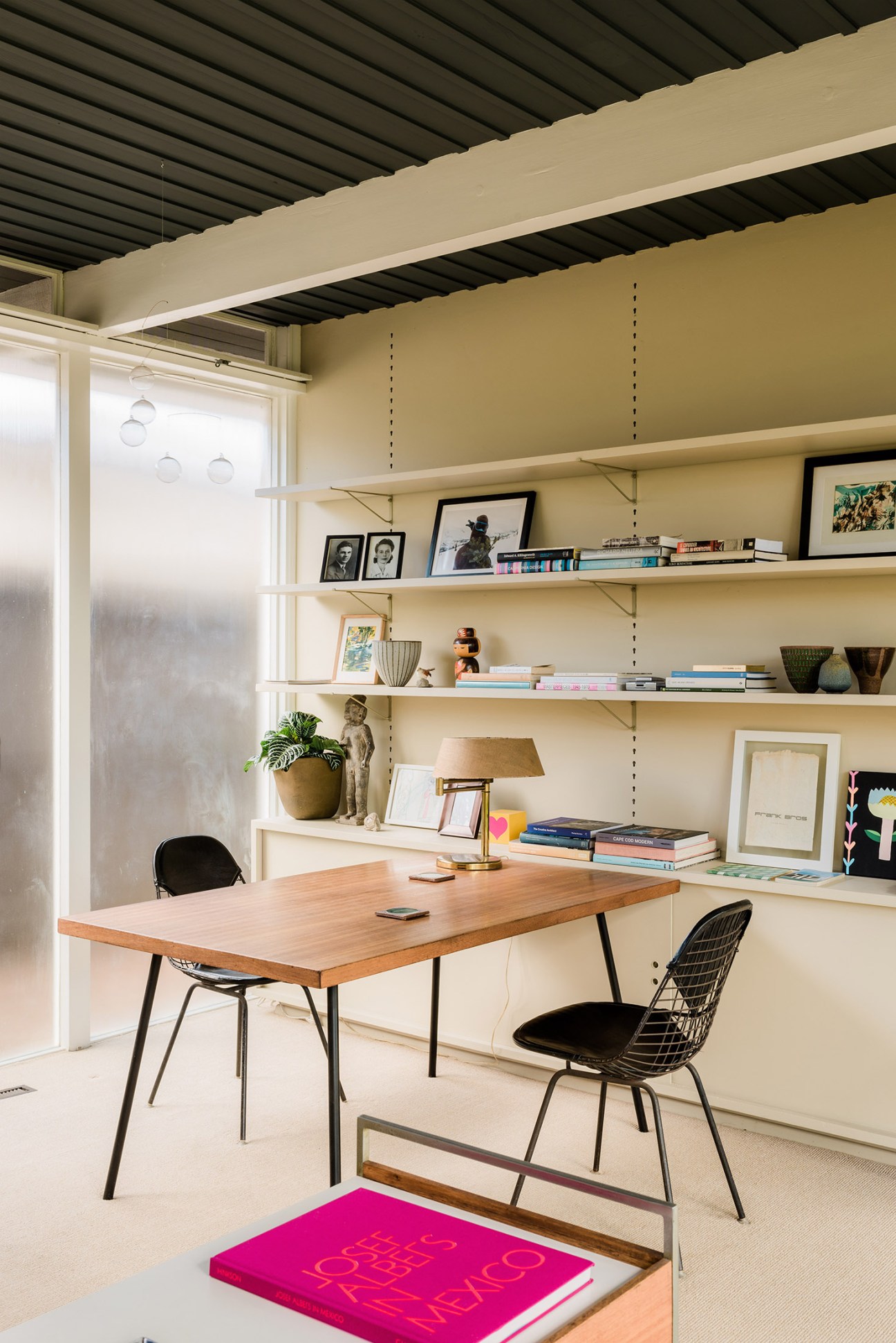 Mid-century modern workspace with wooden desk, metal-legged chairs, yellow walls, and built-in shelving displaying books and decorative objects.