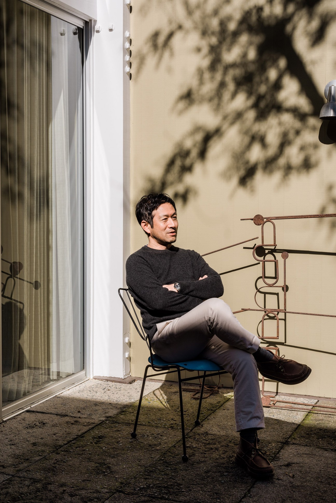 Keiro Koga sits in a mid-century modern chair on the patio of his Killingsworth-designed home, with tree shadows cast on the wall behind him.