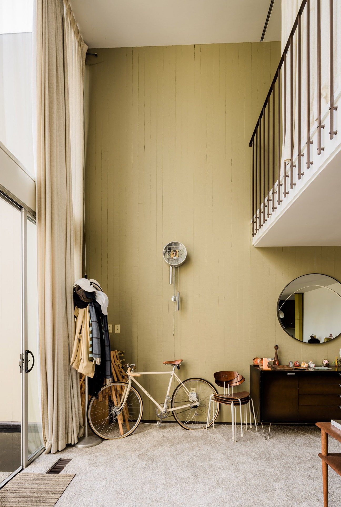 A white Pashley bicycle leans against a yellow-green wall in a modernist living room with a round mirror and wooden furniture.