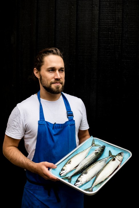 Chef Will Verdino holds a tray of fresh mackerel against a dark wooden backdrop.