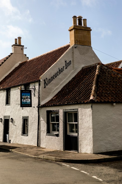 Exterior of the Kinneuchar Inn, a 17th-century whitewashed Scottish building with clay tile roof and tall chimneys.