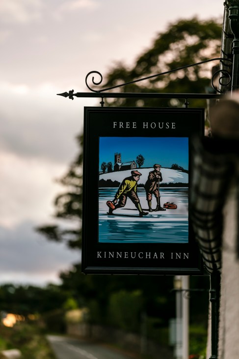 Ornamental pub sign for Kinneuchar Inn featuring illustrated figures on ice with church in background.