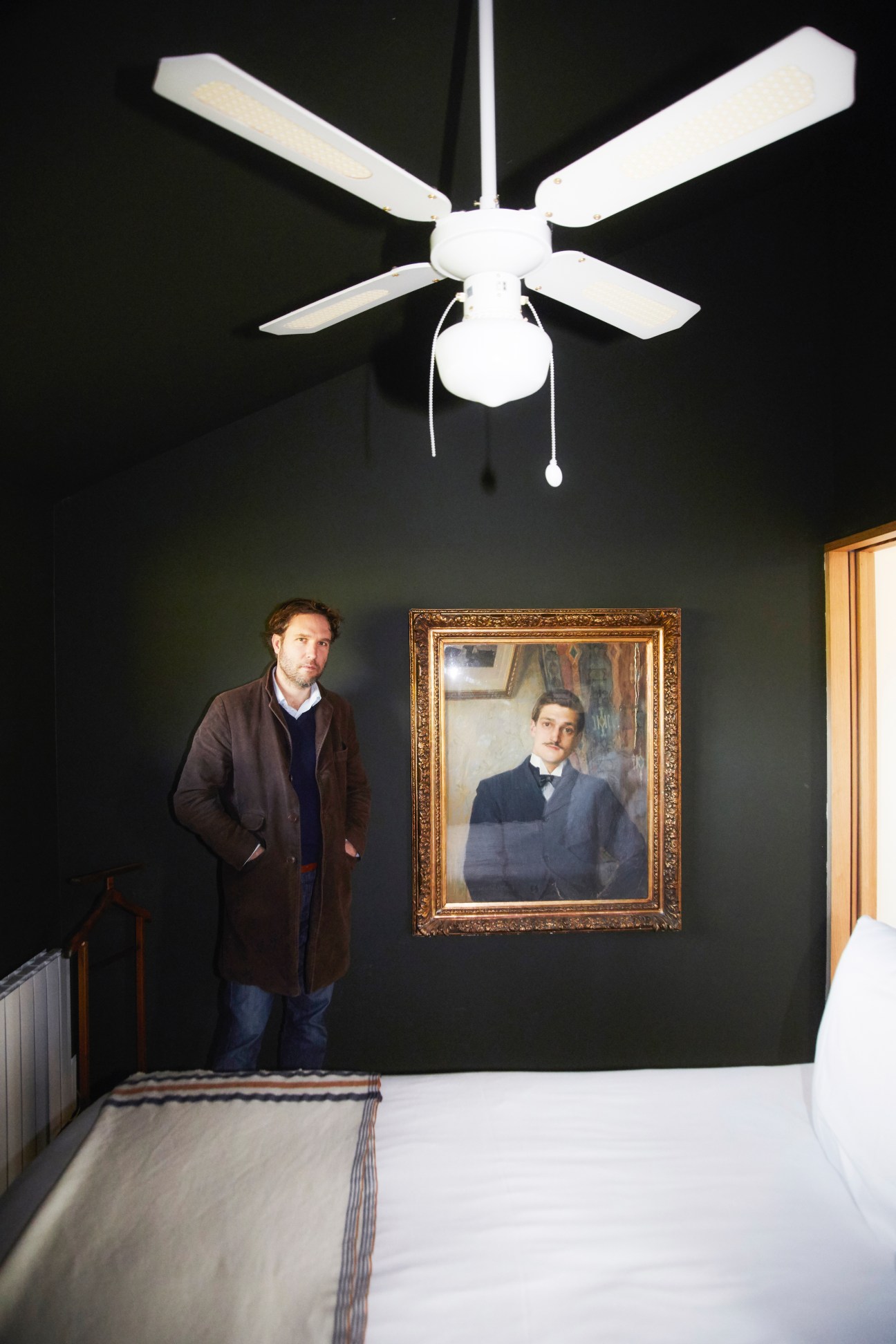 Édouard Daehn stands in a dark bedroom with white ceiling fan and framed portrait on wall