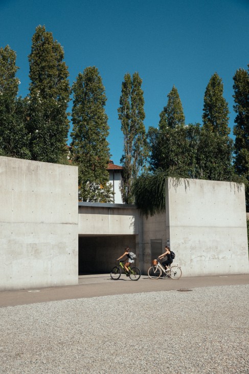 Two cyclists ride past a modern concrete building with tall poplar trees against a blue sky in Locarno.