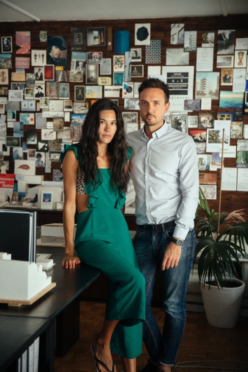 Architect Matteo Inches and his wife Nastasja Inches-Geleta pose in front of a wall covered with framed artwork and photographs.