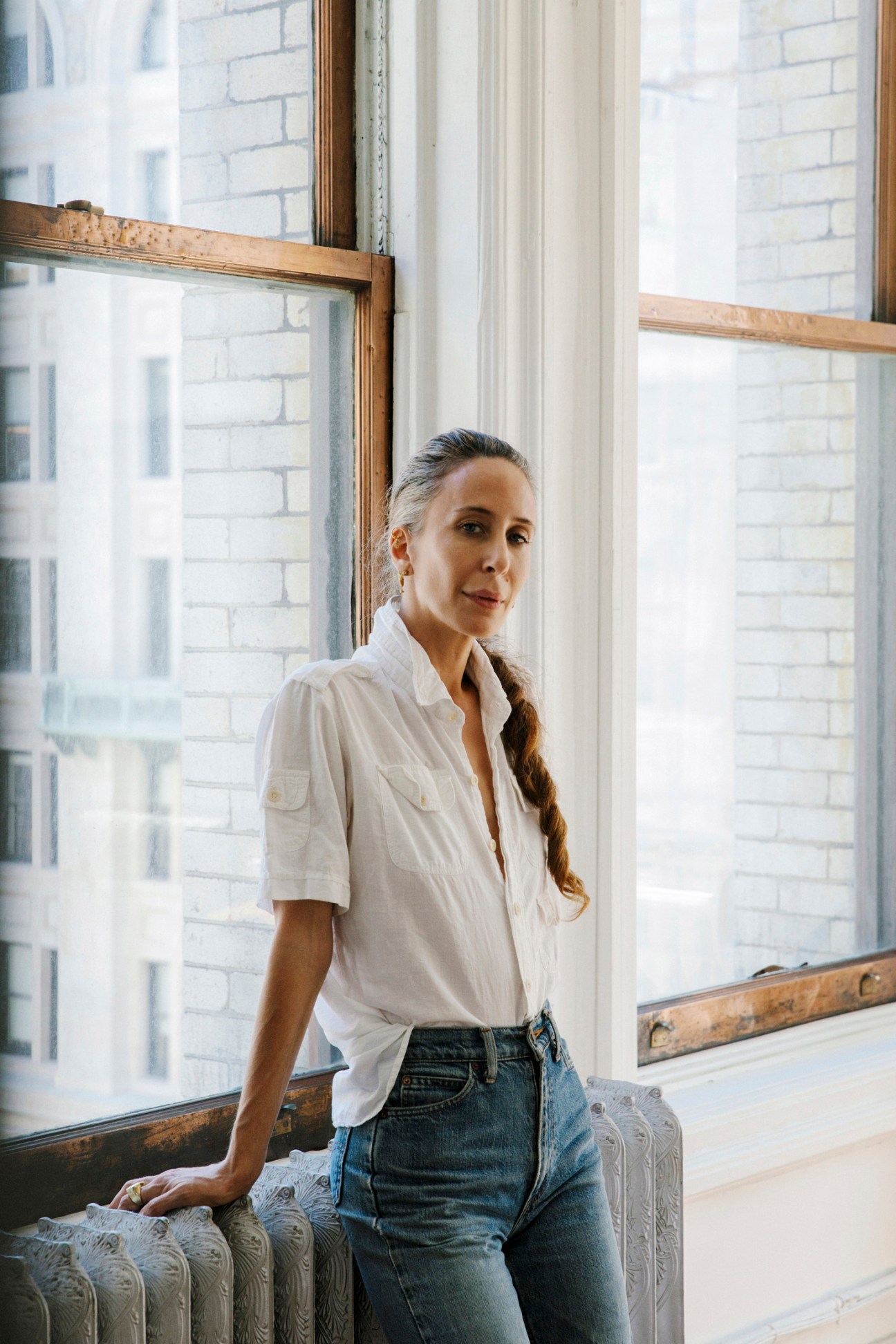 Designer Mara Hoffman standing by a window in her studio, wearing a white shirt and jeans.