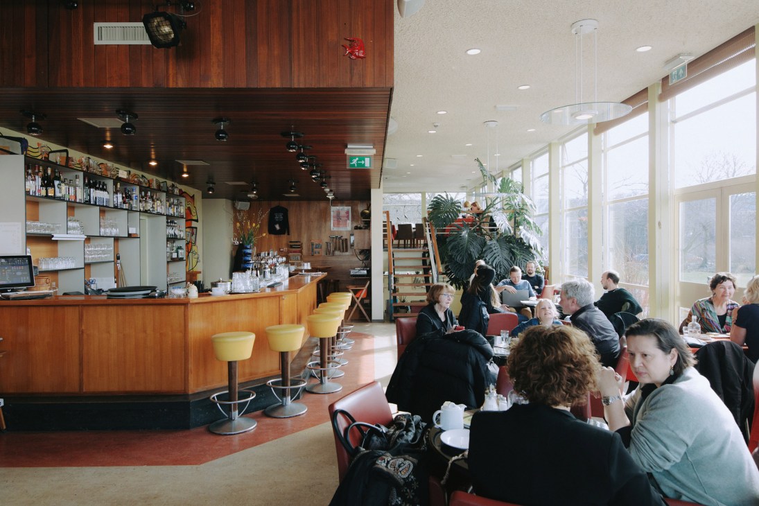 Interior of Pension Homeland café with wooden bar, yellow stools, and diners seated at tables near windows.