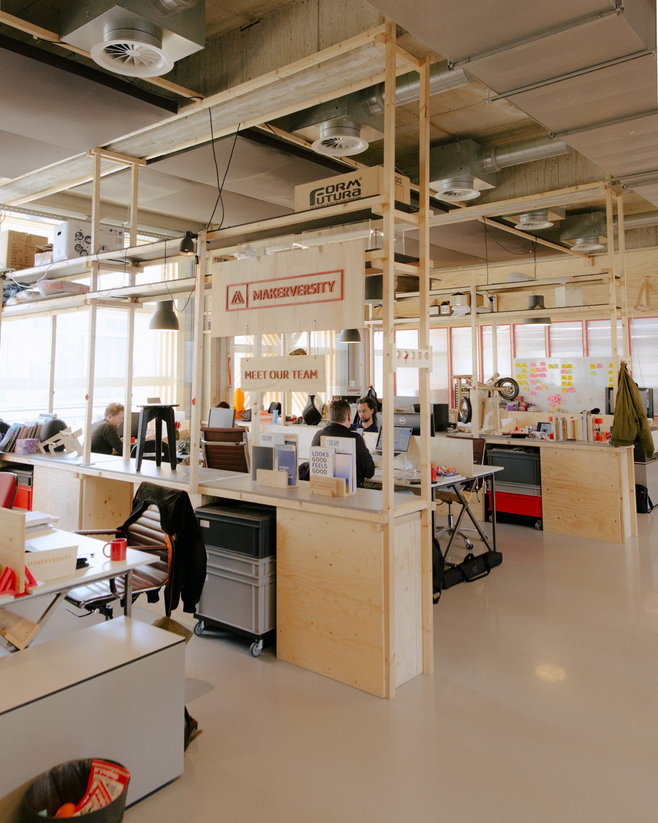 Interior of Makerversity workspace with wooden modular partitions, people working at desks, and signage reading 'Meet Our Team'.