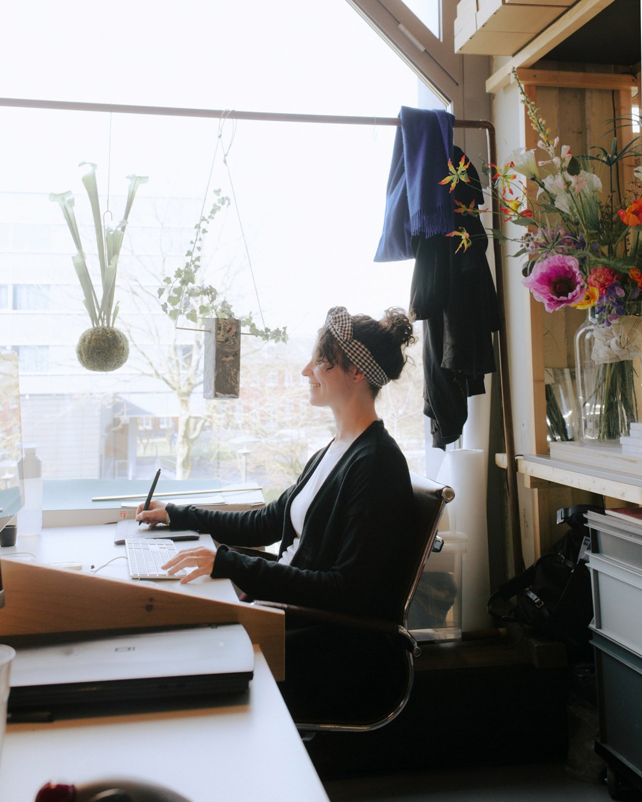 Woman working at a desk by a window in a bright workspace with hanging plants and flowers.