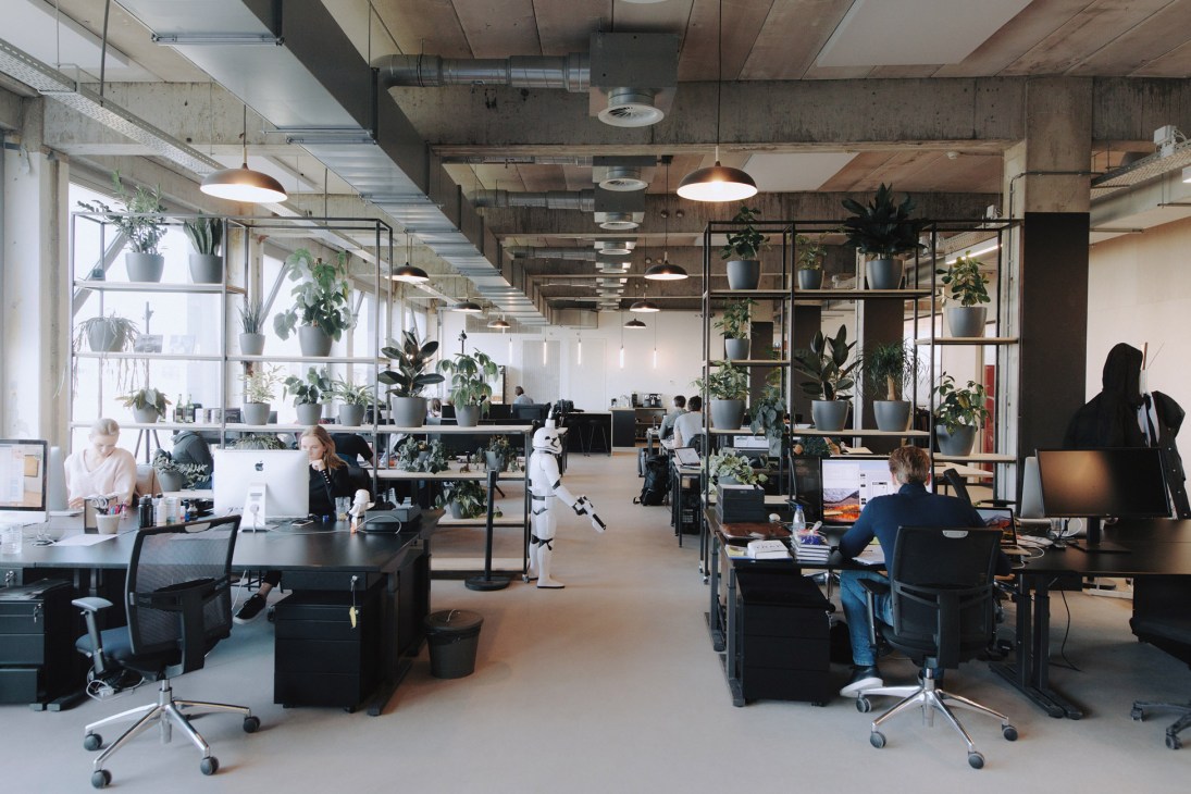 Modern open-plan office workspace with exposed concrete ceiling, pendant lighting, desk stations, and potted plants throughout.