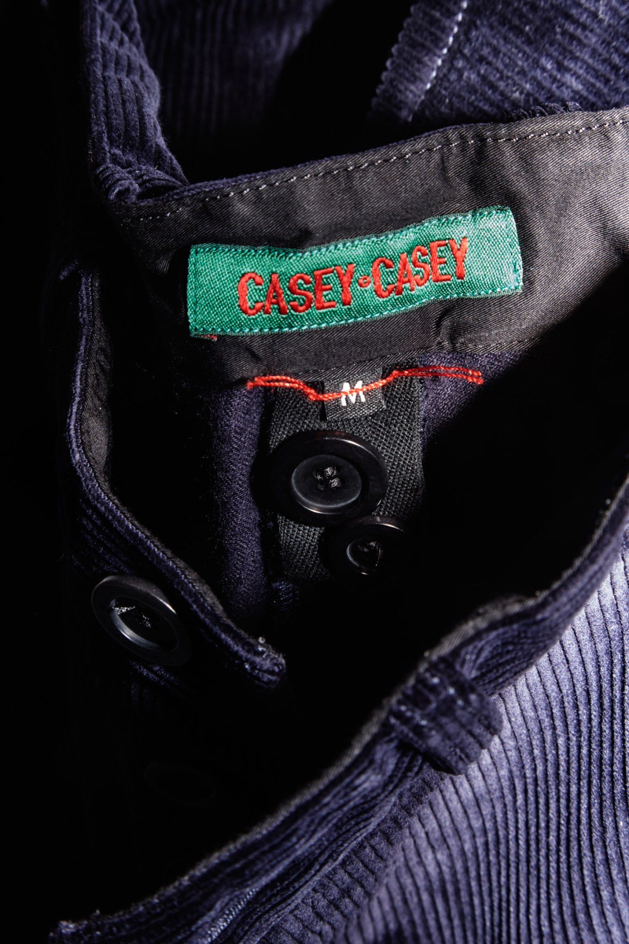 Close-up of a Casey Casey clothing label with red text on green and gray fabric with black buttons.