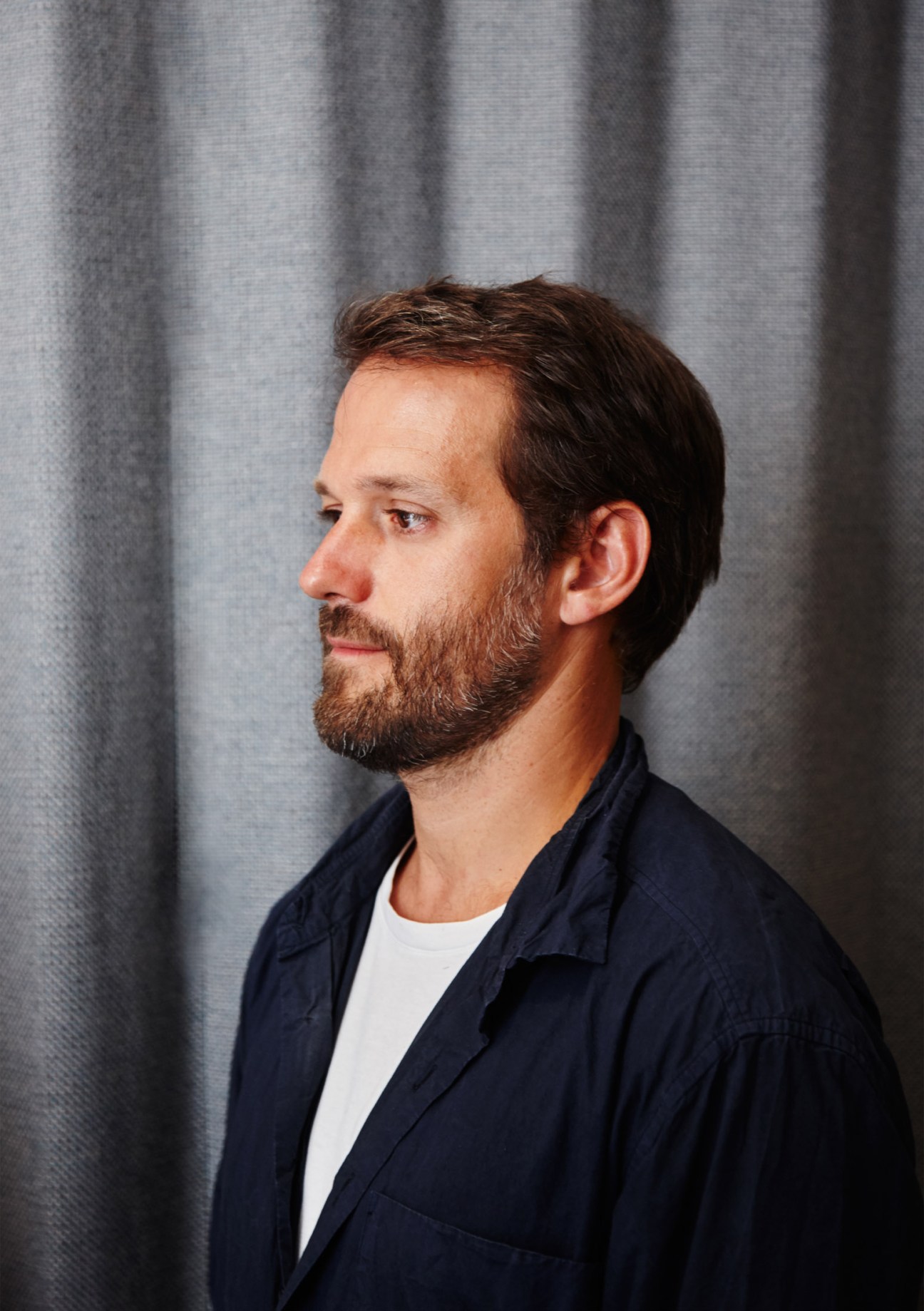 Man in profile wearing dark navy shirt and white t-shirt against gray curtain backdrop.