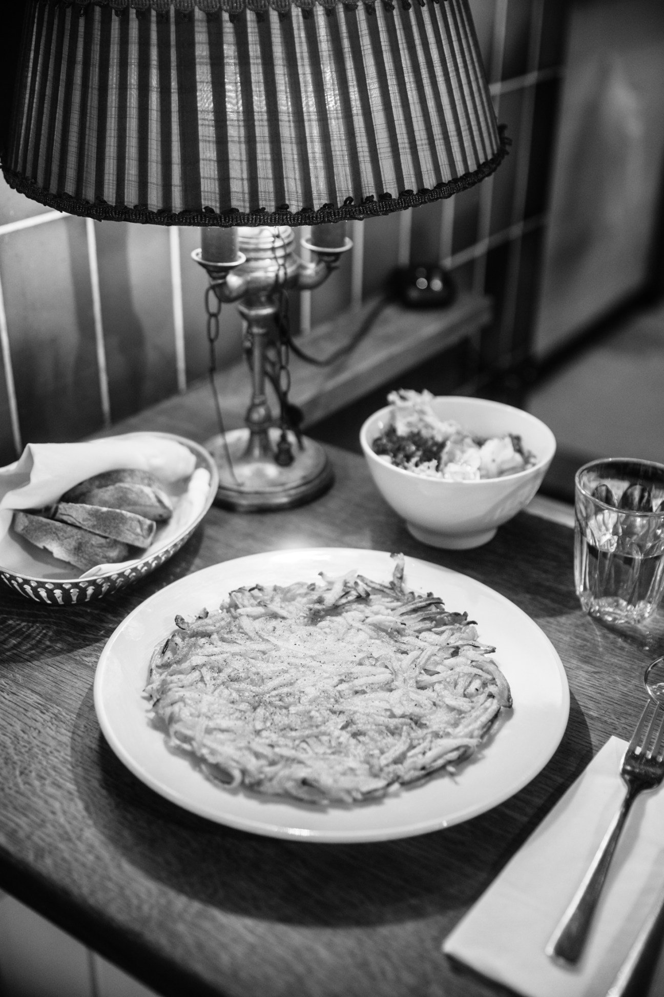 Black and white photo of pasta dish with lamp, bread basket, salad bowl and glass on wooden table