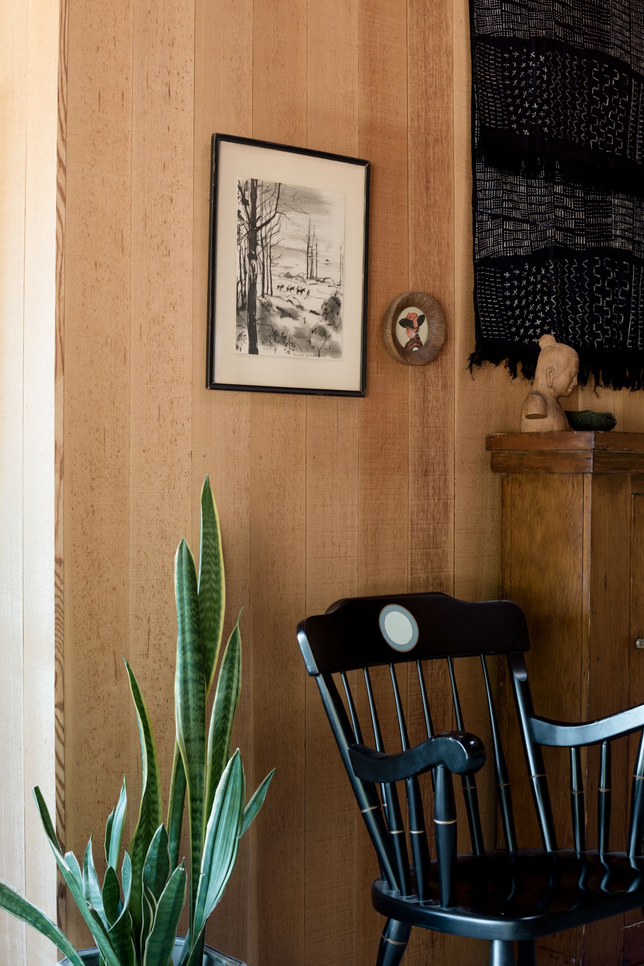 Wooden interior wall with framed landscape drawing, black chair, potted plant, and decorative objects on shelf.