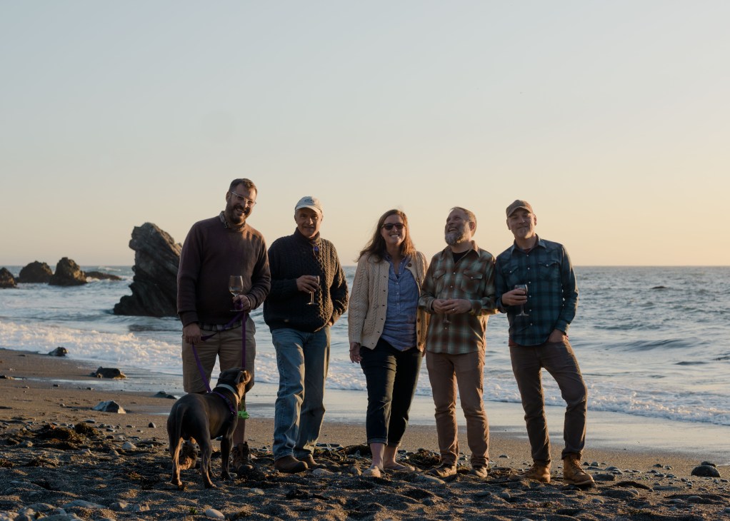 Six Sea Ranch residents and a dog gather on a pebble beach at sunset with ocean waves behind them.
