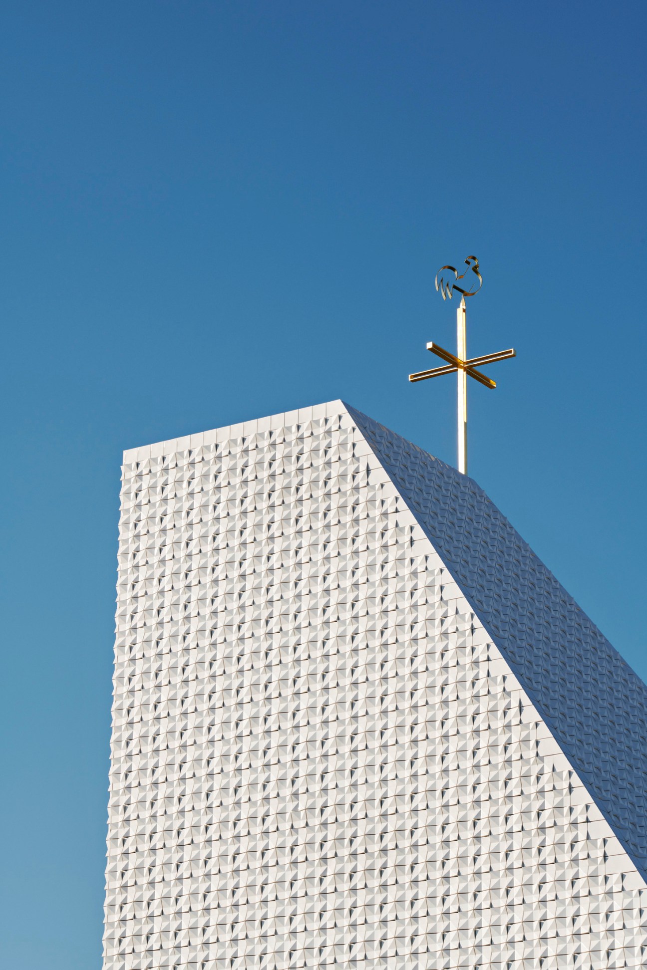 A tall white church wall with a cross and weathervane on top against a blue sky.