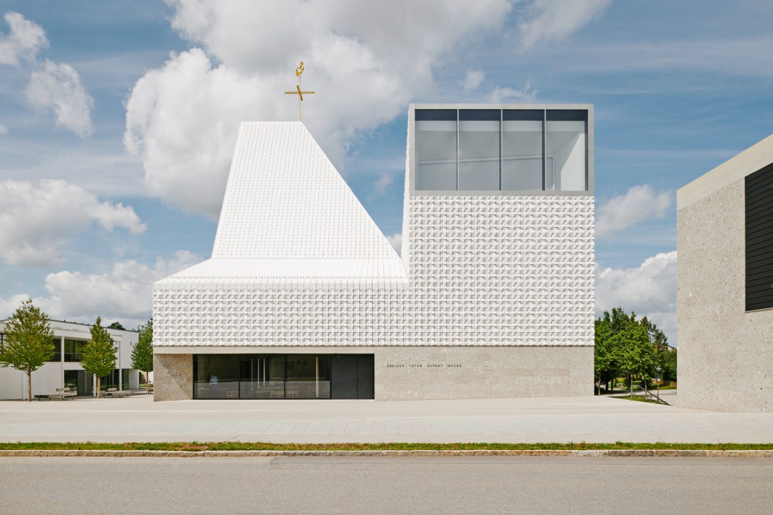 Church of the Blessed Father Rupert Mayer in Poing, Germany, featuring white ceramic tiles on upper section and natural stone base under blue sky.