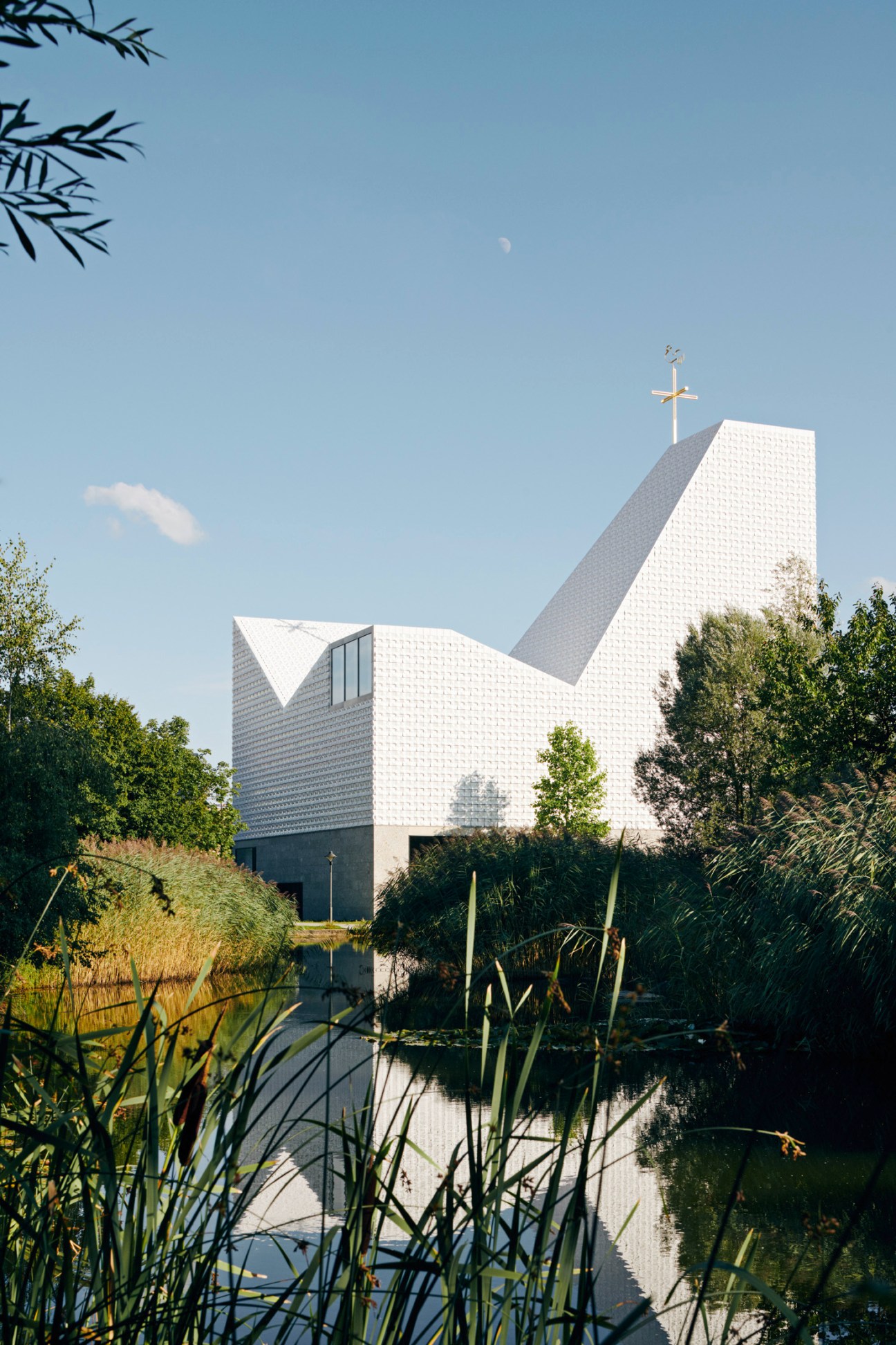 Church of the Blessed Father Rupert Mayer in Poing, Germany, with white ceramic tile roof and cross, surrounded by water and vegetation.