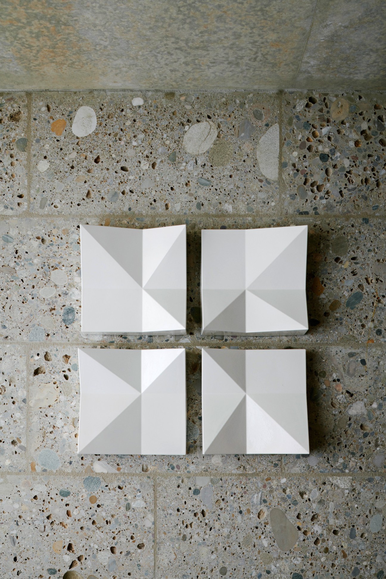 Four white ceramic tiles with geometric folded patterns arranged on a textured concrete surface.