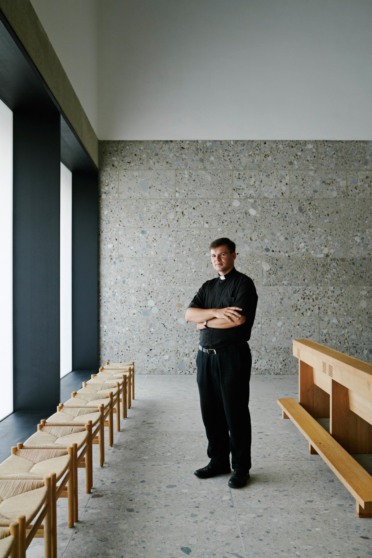 A priest in black robes stands in the minimalist interior of Church of the Blessed Father Rupert Mayer, with wooden pews and concrete walls.