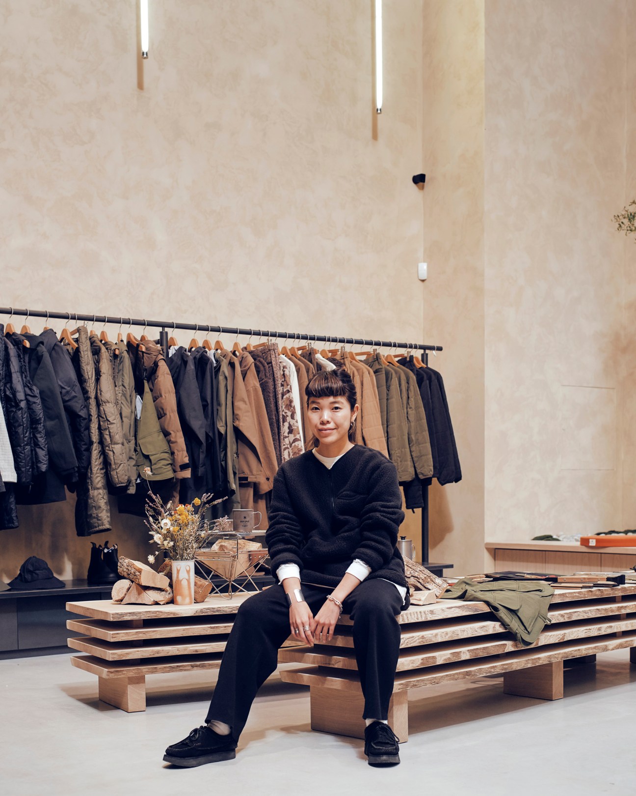 Lisa Yamai sitting on wooden bench in Snow Peak store wearing black V-neck fleece with clothing racks behind her