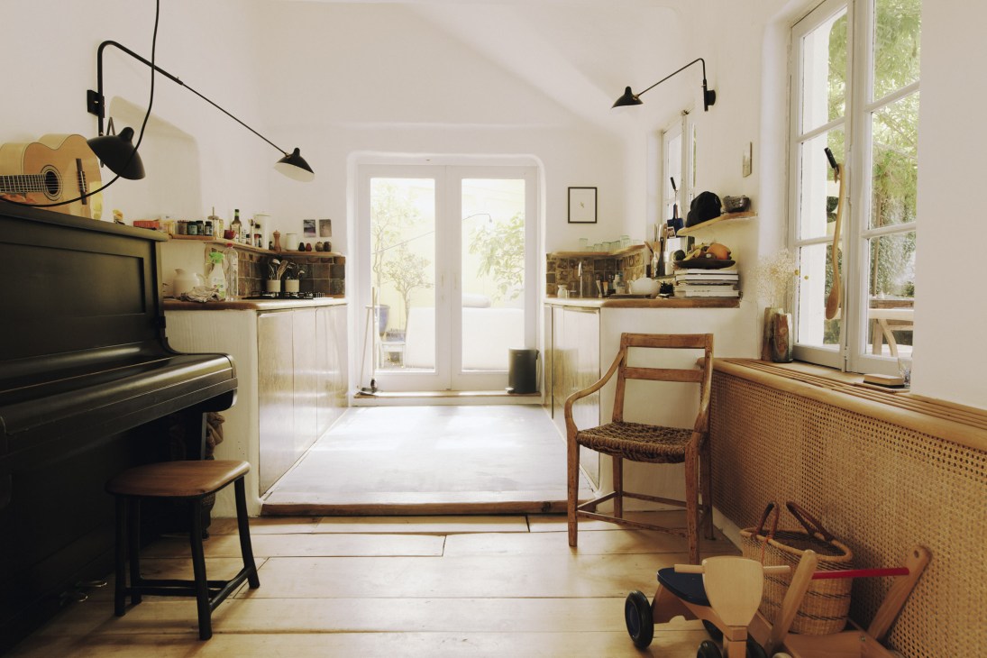 Bright kitchen with Serge Mouille pendant lights, white plaster walls, wooden shelving, brass cabinets, and garden views through French doors.
