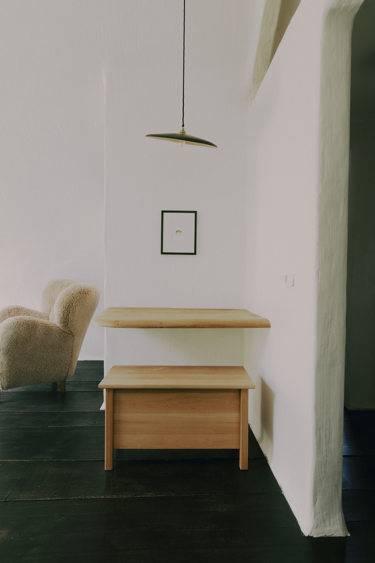 Minimalist bedroom with white walls, wooden furniture, Serge Mouille pendant light, and cream upholstered chair.