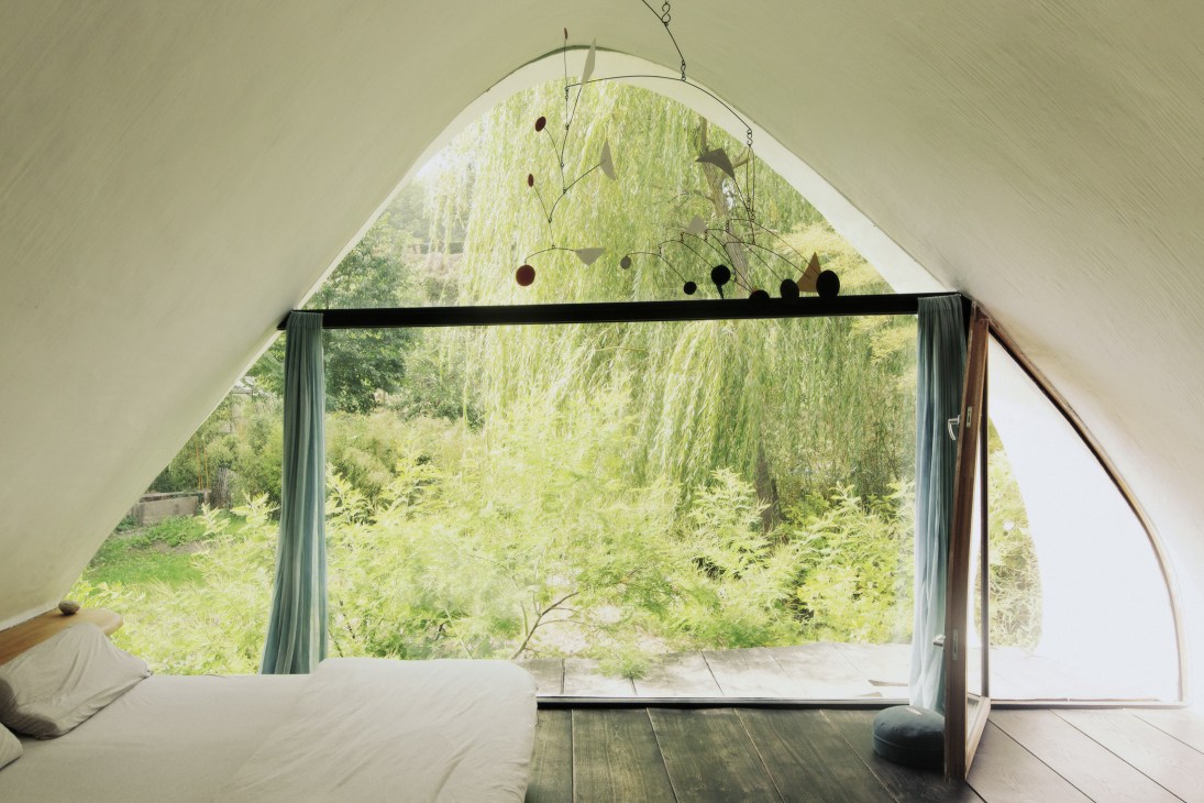 Bedroom with curved window overlooking garden, featuring Calder-inspired mobile and white walls.