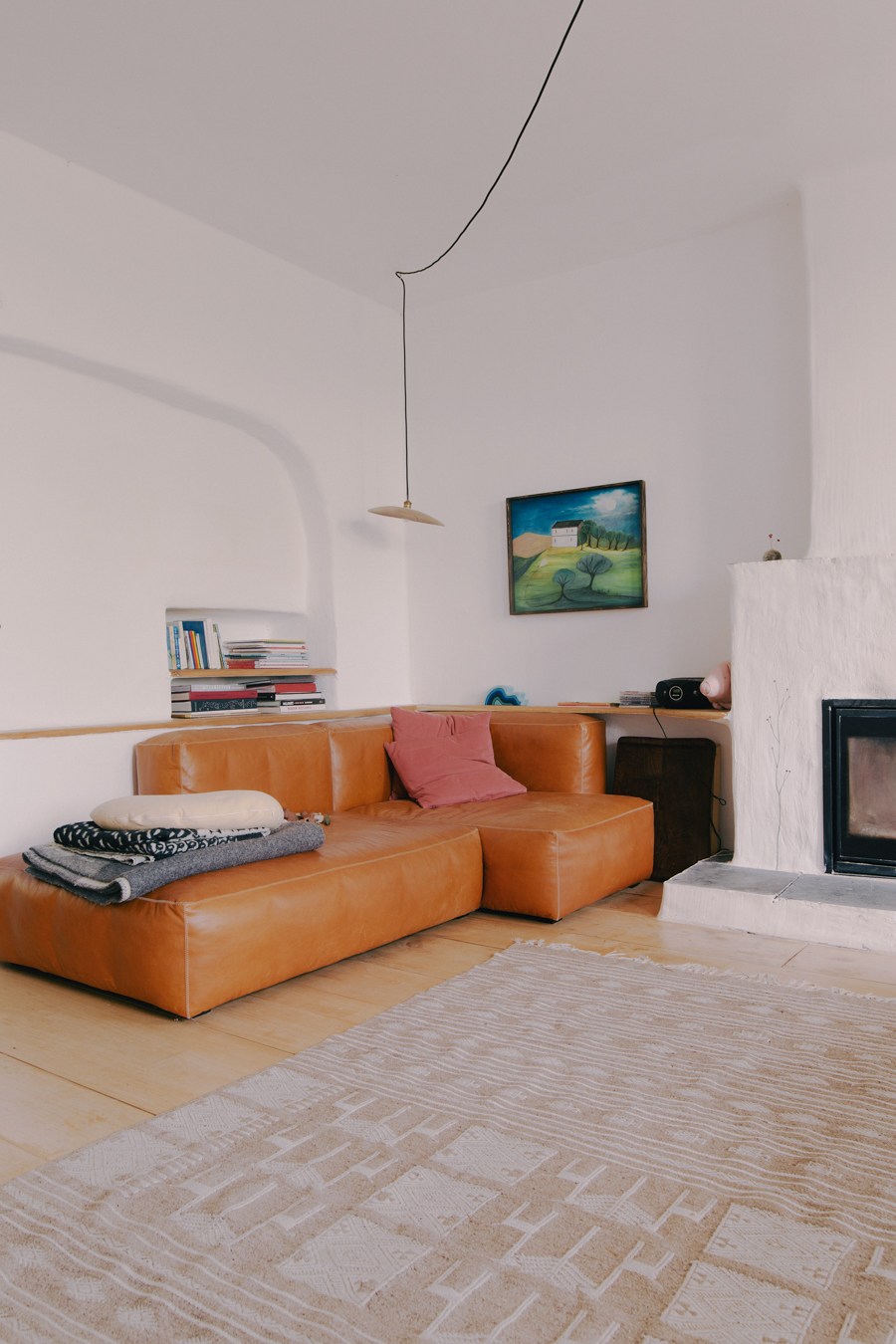 A living room with white plaster walls, orange leather sofa, pendant lighting, and a painting by Valentin Loellmann's grandmother on the wall.