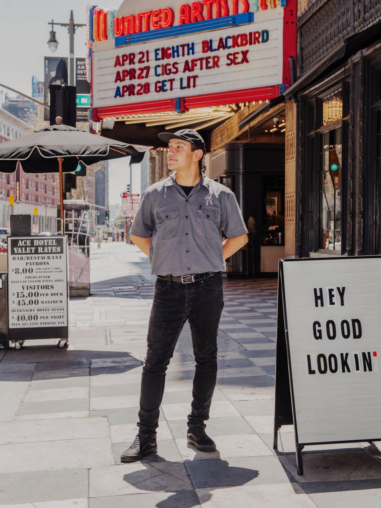 Man in gray shirt and cap standing outside United Artists theater with marquee in background.