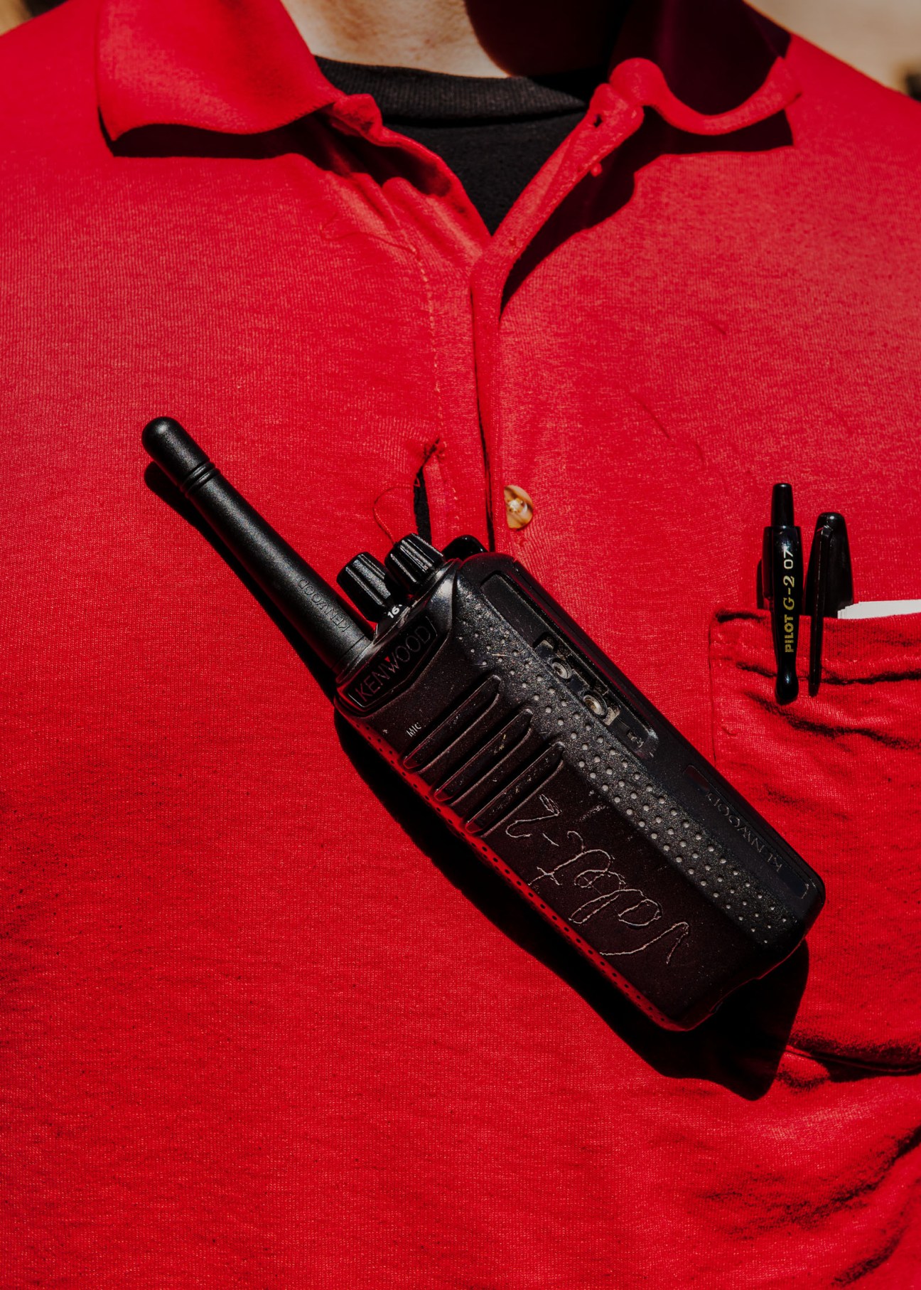 A valet in a red uniform holds a walkie-talkie and pen against their chest.