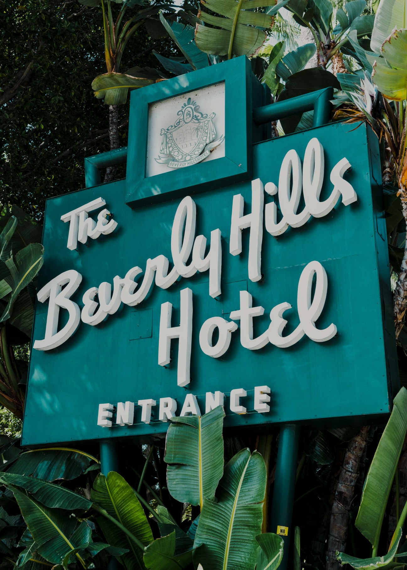 Turquoise entrance sign for The Beverly Hills Hotel surrounded by tropical foliage.