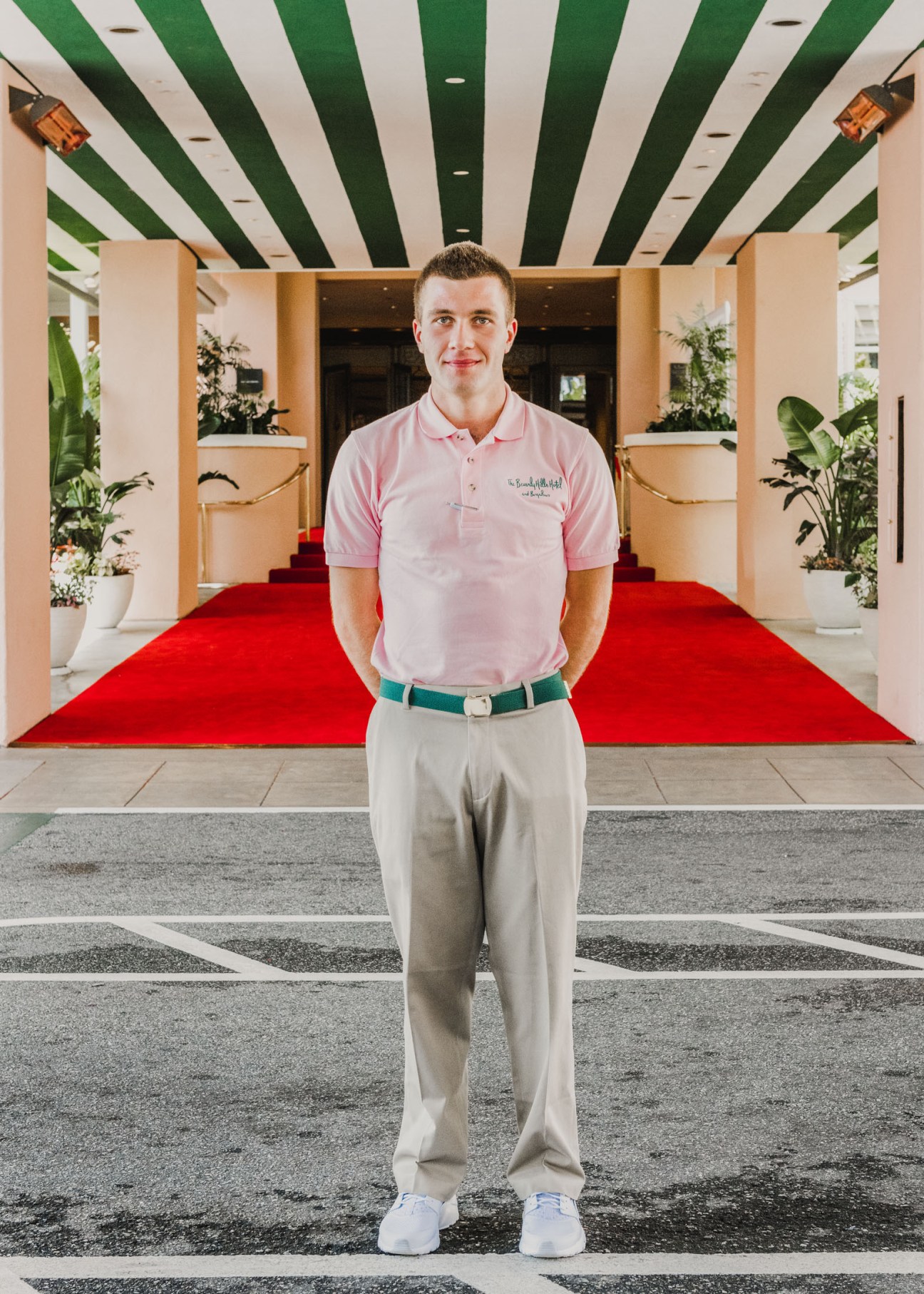 Valet parker in pink polo shirt and khakis standing in hotel entrance with red carpet and striped ceiling