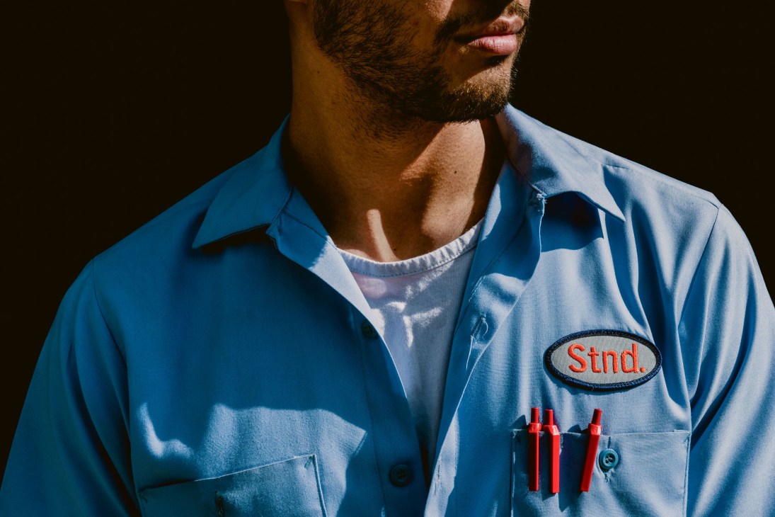 Man wearing blue valet uniform with name patch and red pens in pocket against dark background.