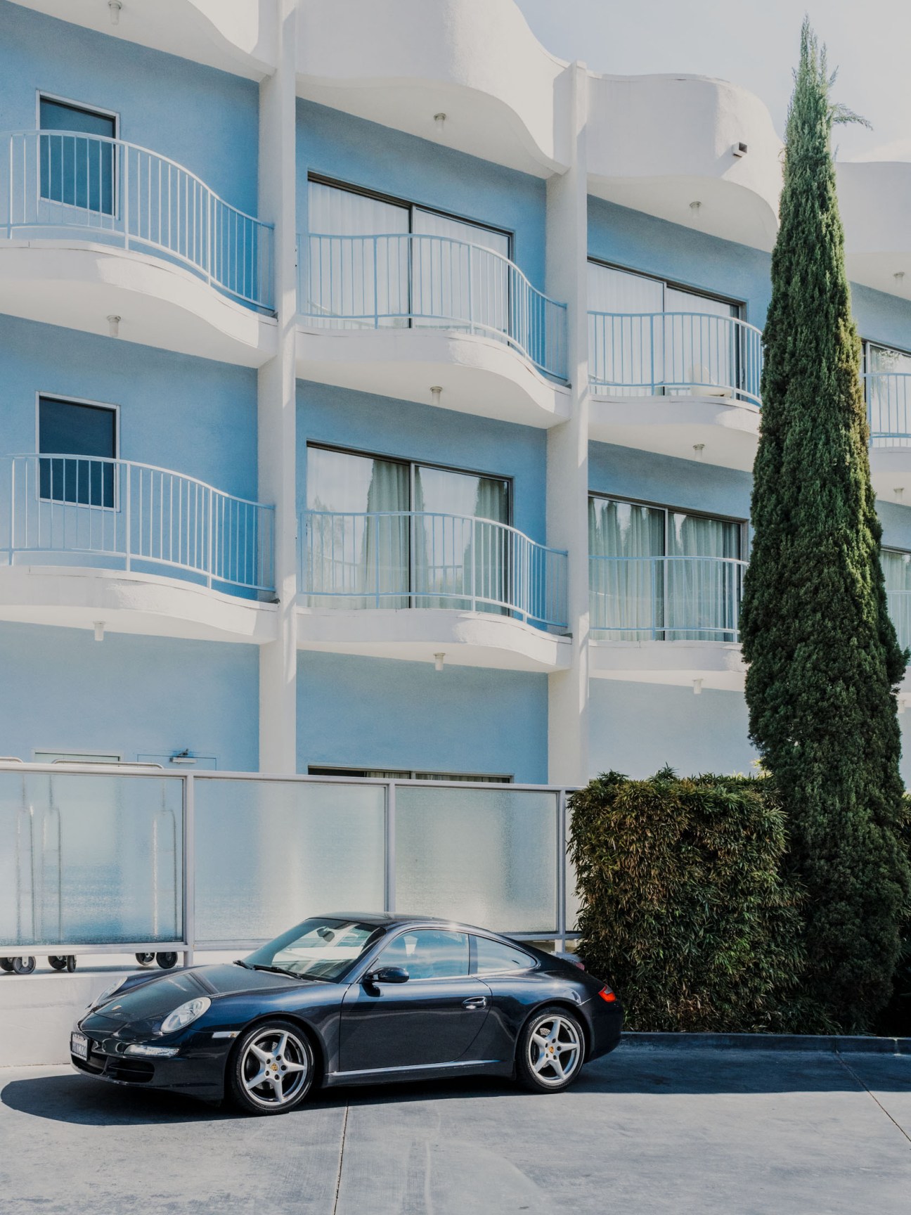 Black Porsche 911 parked in front of modern blue and white building with balconies and cypress tree.