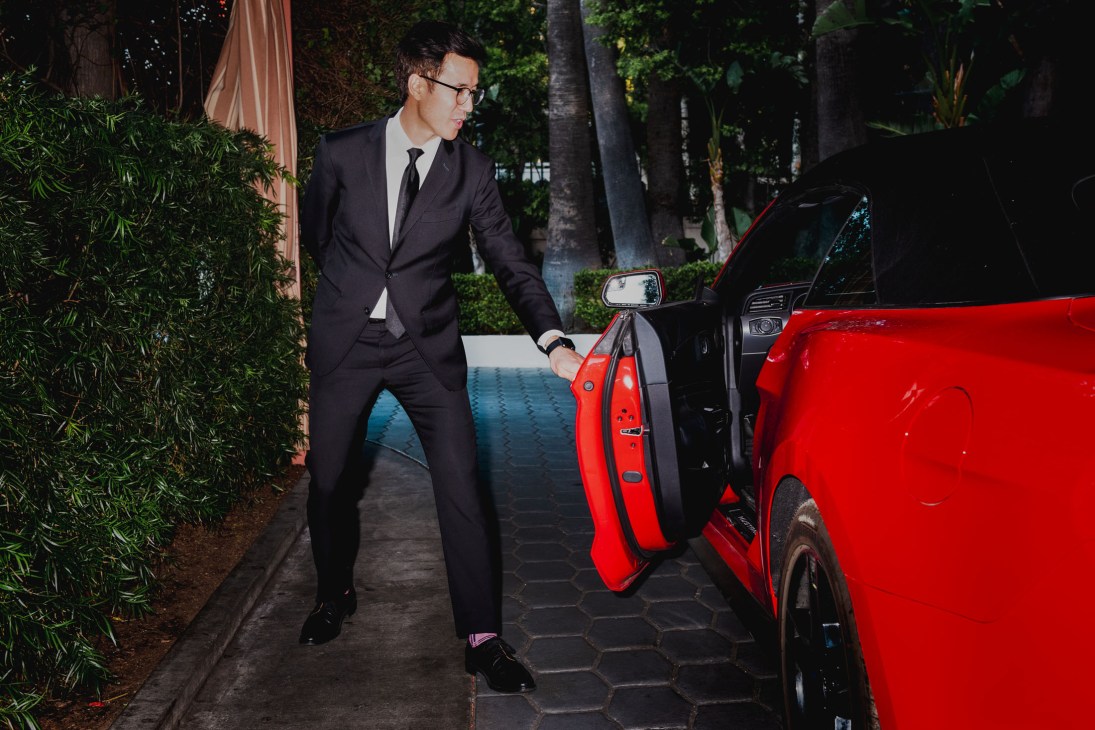 A valet in a dark suit and tie stands beside a red car, reaching for the door handle in a professional pose.