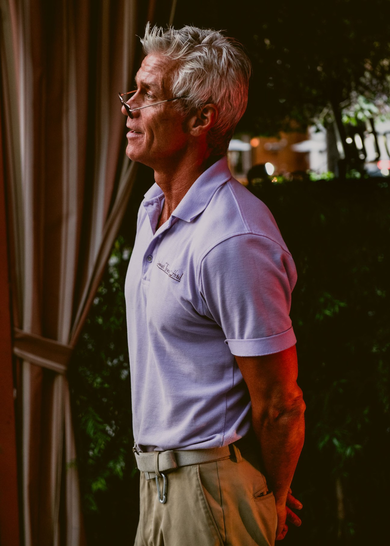 An older man wearing glasses and a lavender polo shirt stands in profile against an evening backdrop.